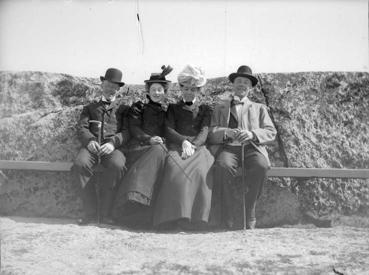 Svartvitt foto. Fyra hattklädda personer, två män och två kvinnor sitter på en bänk framför en klipphäll.