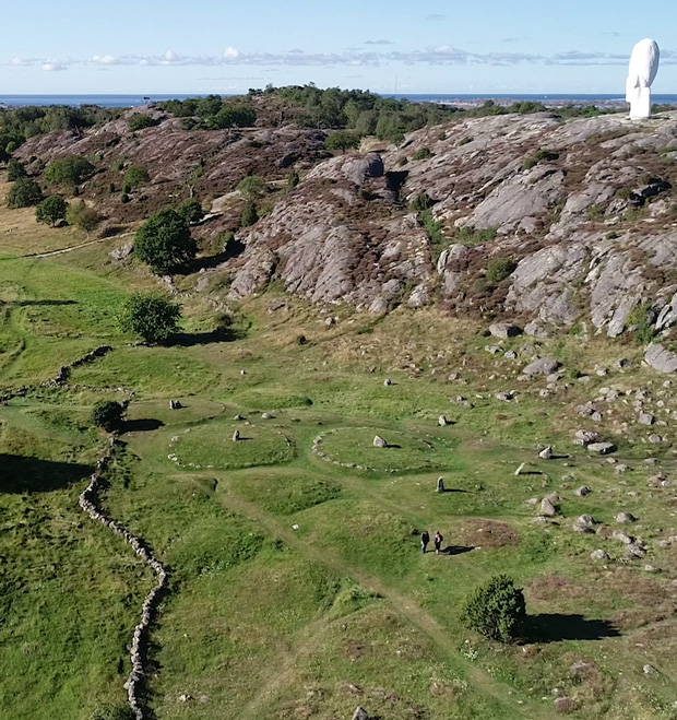 Foto. Vy över ett kustlandskap med fyra cirkelformade gräs-kullar. På mitten av kullarna står en meterhög sten av granit. 