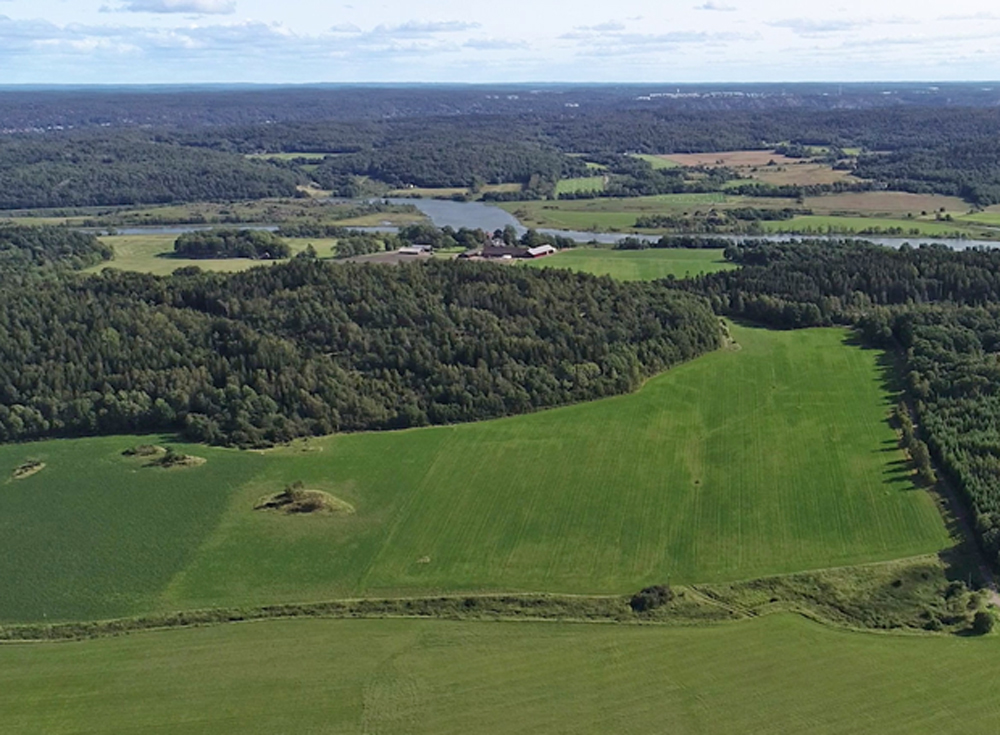 Färgfoto från luften på åkermark omgiven av skogsklädda berg. I bakgrunden slingrar sig älven.