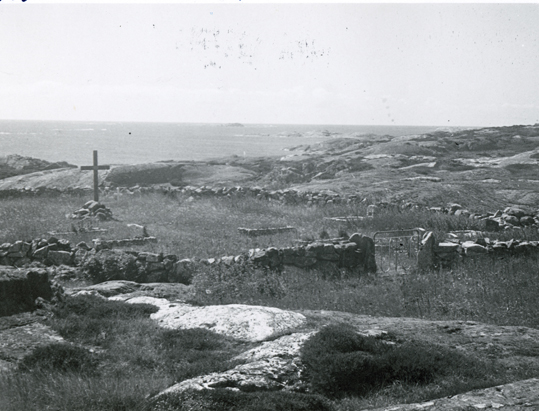 Svartvitt foto av ett kalt bergigt kustlandskap med ett inhägnat och gräsbevuxet område, i vars mitt det finns ett stående kors.