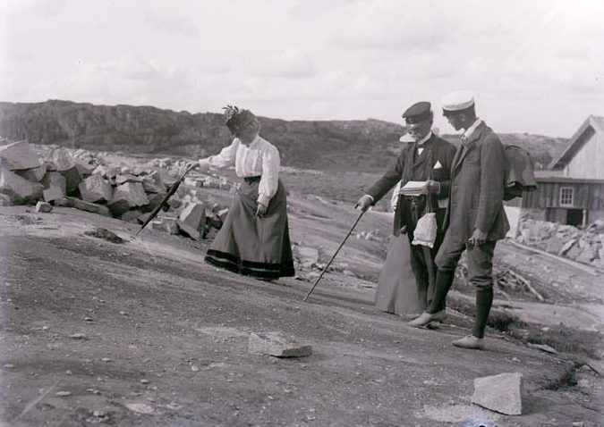 Svarvitt foto. En medelålders kvinna i hatt, blus och lång kjol pekar på en berghäll med en käpp. Bredvid står två män.