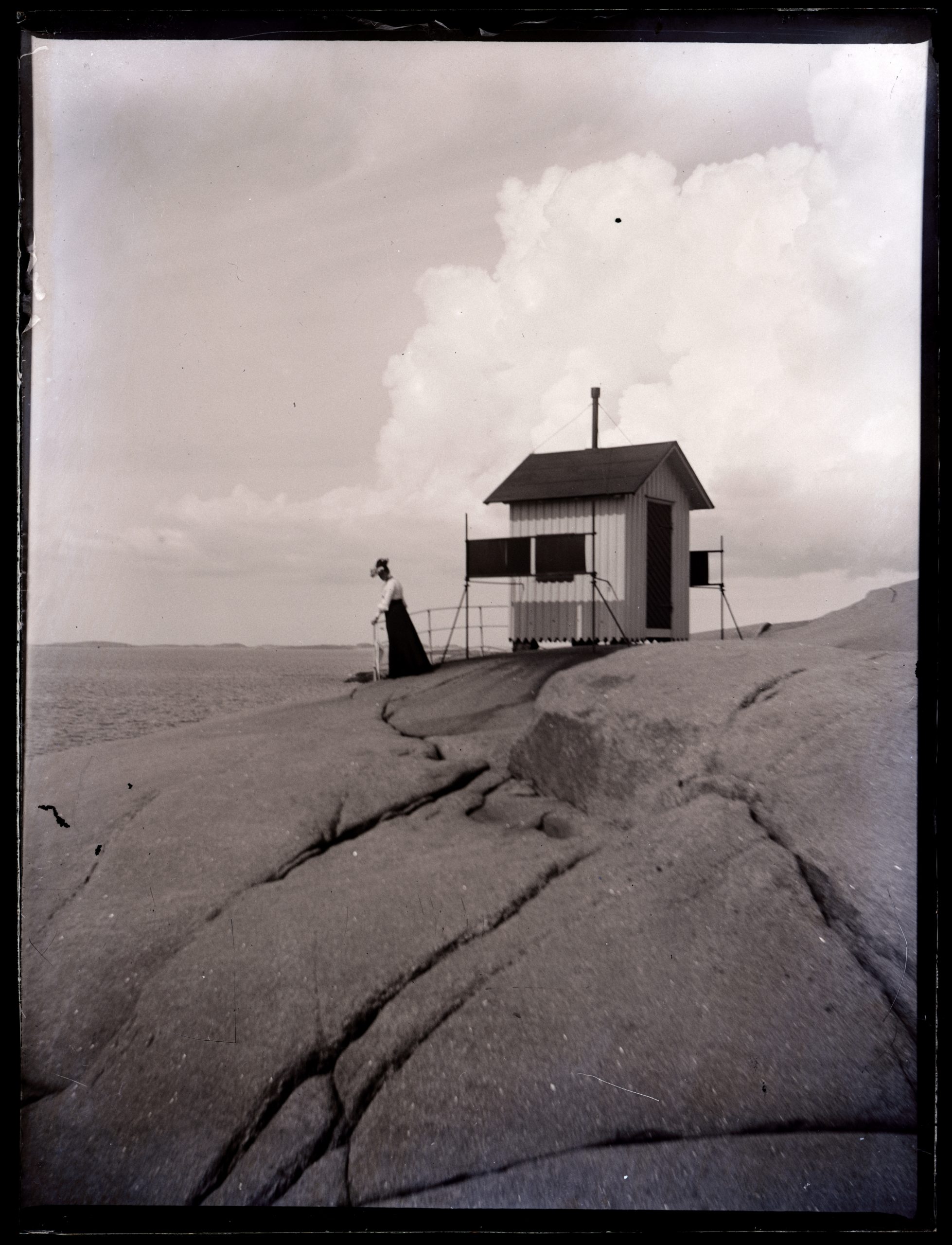 Svartvitt foto. På en hög klippa står en kvinna vid en liten ljusmålad byggnad och ser ut över havet.