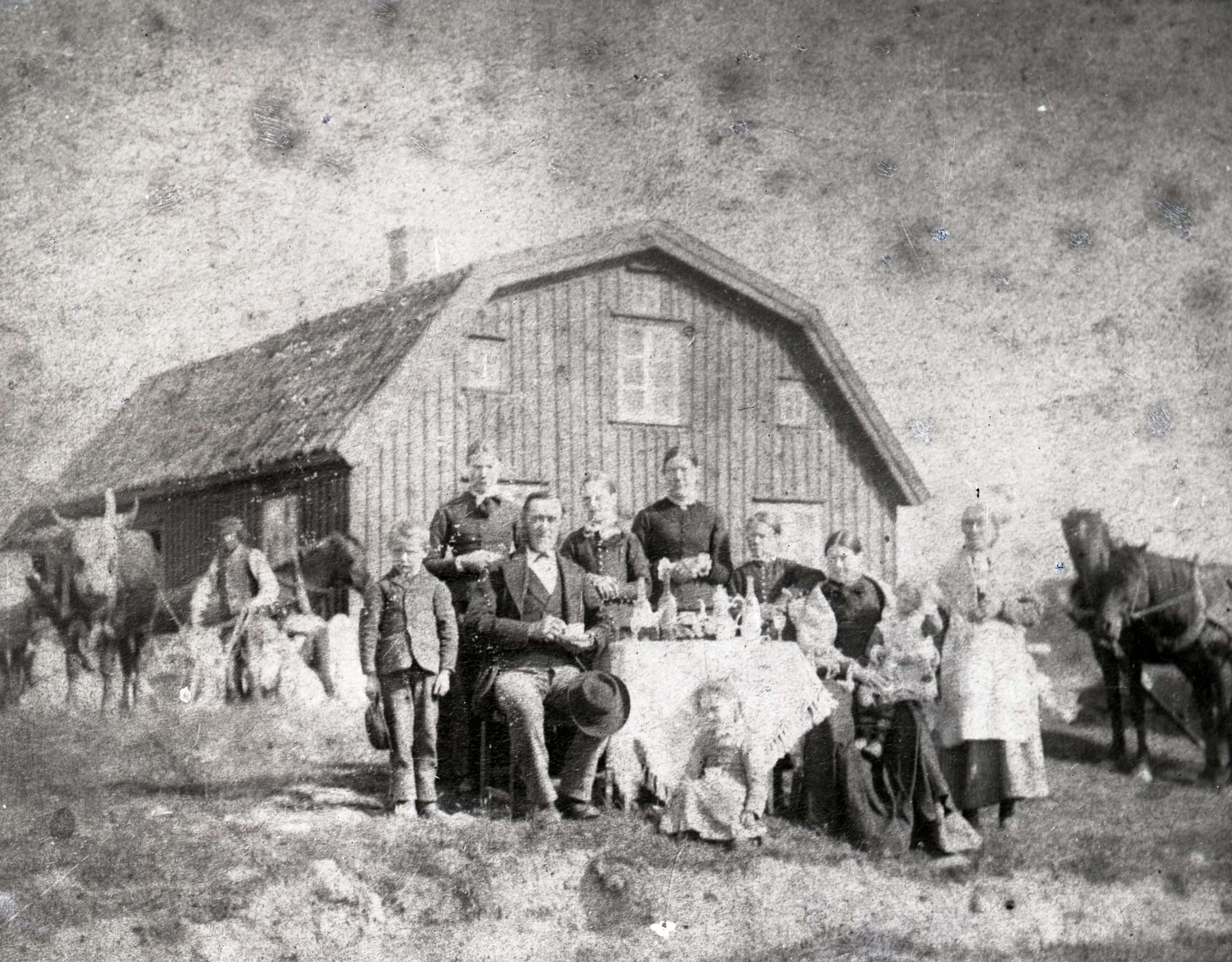 Svartvitt foto. En familj sitter och står vid ett dukat bord framför gaveln av ett botadshus. Även tjänstefolk och boskap är med på bilden.