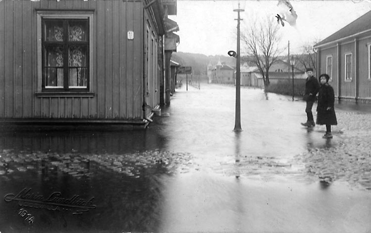Svartvitt foto. En översvämmad stadsgata flankerad av envånings trähus, vars stengrund står i vatten. Två barn står på gatan och tittar nyfiket in i kameran.