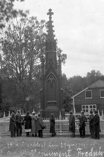 Svartvitt foto med personer vid ett högt tornprytt minnesmonument omgivet av ett gjutjärnsstaket.