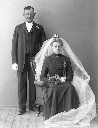 Svartvitt foto av brudpar. Den sittande bruden har mörk klänning med hög hals och snävt liv. Vit lång brudslöja med liten myrtenkrona. I handen en brudbukett.