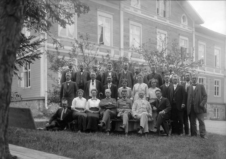 Svartvitt foto men en grupp kvinnor och män som sitter och står framför en stor byggnad.