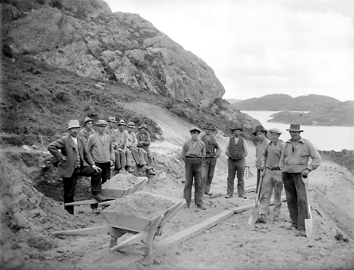 Svartvitt foto där vägarbetare står vid skottkärror av trä lastade med vägmaterial. I bakgrunden skymtar Gullmarsfjorden.