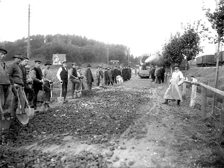 Svartvitt foto där en rad män med spadar står vid en påbörjad vägsträcka. En ångvält sysn i bakgrunden.