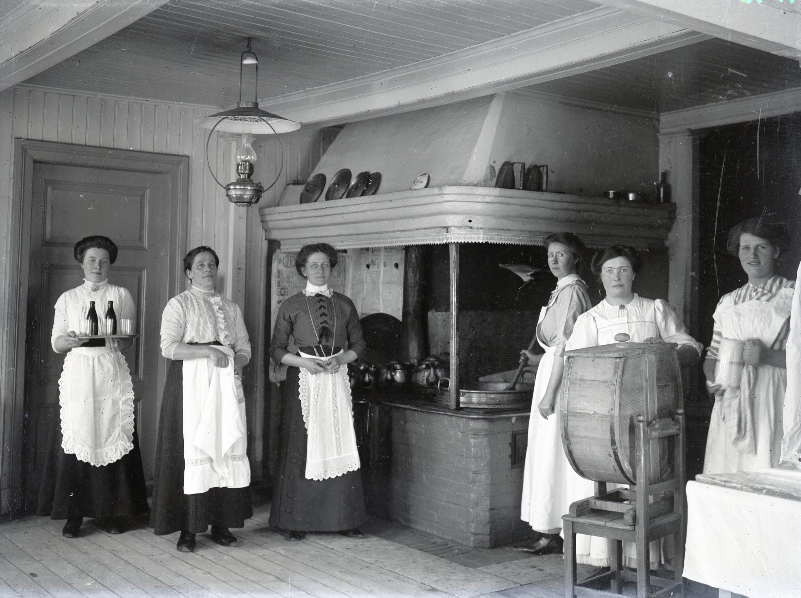 Svartvitt foto av köksinteiör. Vid en stor murad spis med kåpa står sex kvinnor i vita förkläden.