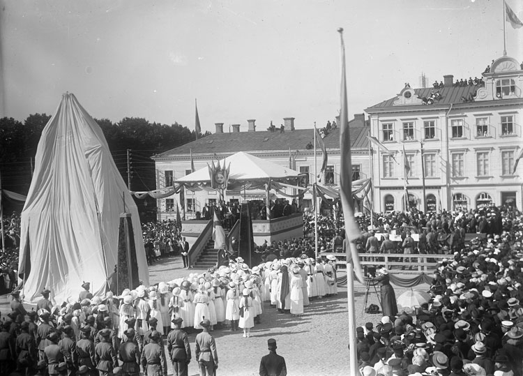 Svartvitt foto. Ett torg fyllt av ett hundratal personer. I mitten av torget finns en upphöjd scen och ett sju meter högt föremål som är dolt av ett skynke.