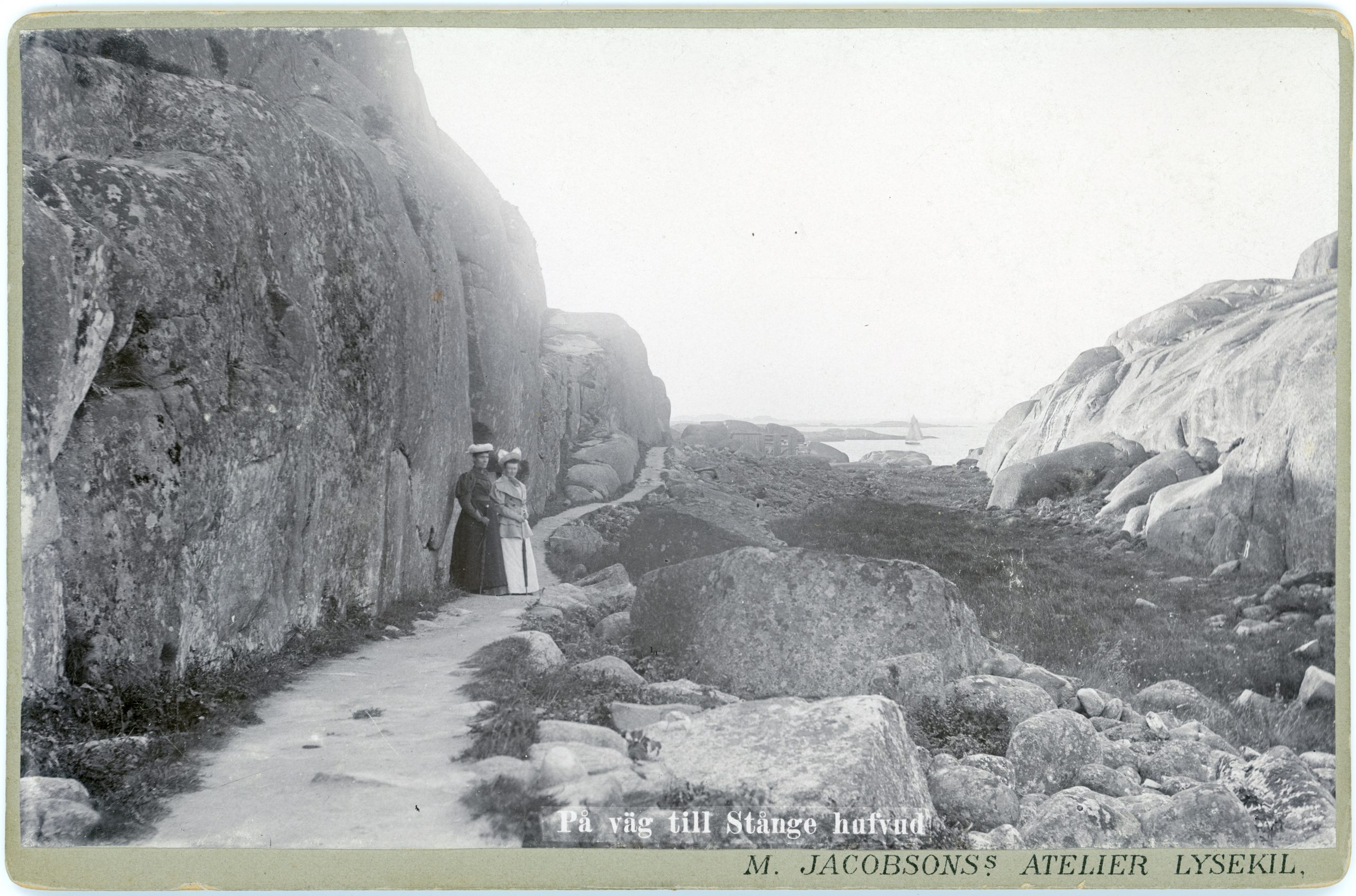 Svartvitt foto. Två kvinnor i hatt och långa dräkter promenerar invid ett högt berg.