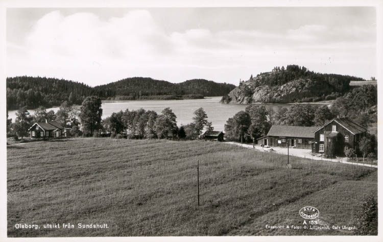 Svartvitt foto visar åker och några hus. I bakgrunden en sjö och skogsklädda bergskullar.