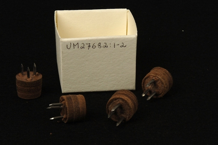 Färgfoto på fyra små runda föremål med metallpiggar samt tillhörande låda.