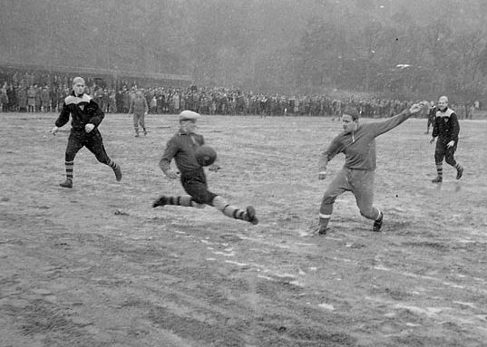 Svartvitt foto med spelare i full fart på en fotbollsplan. Åskådare syns i bakgrunden.