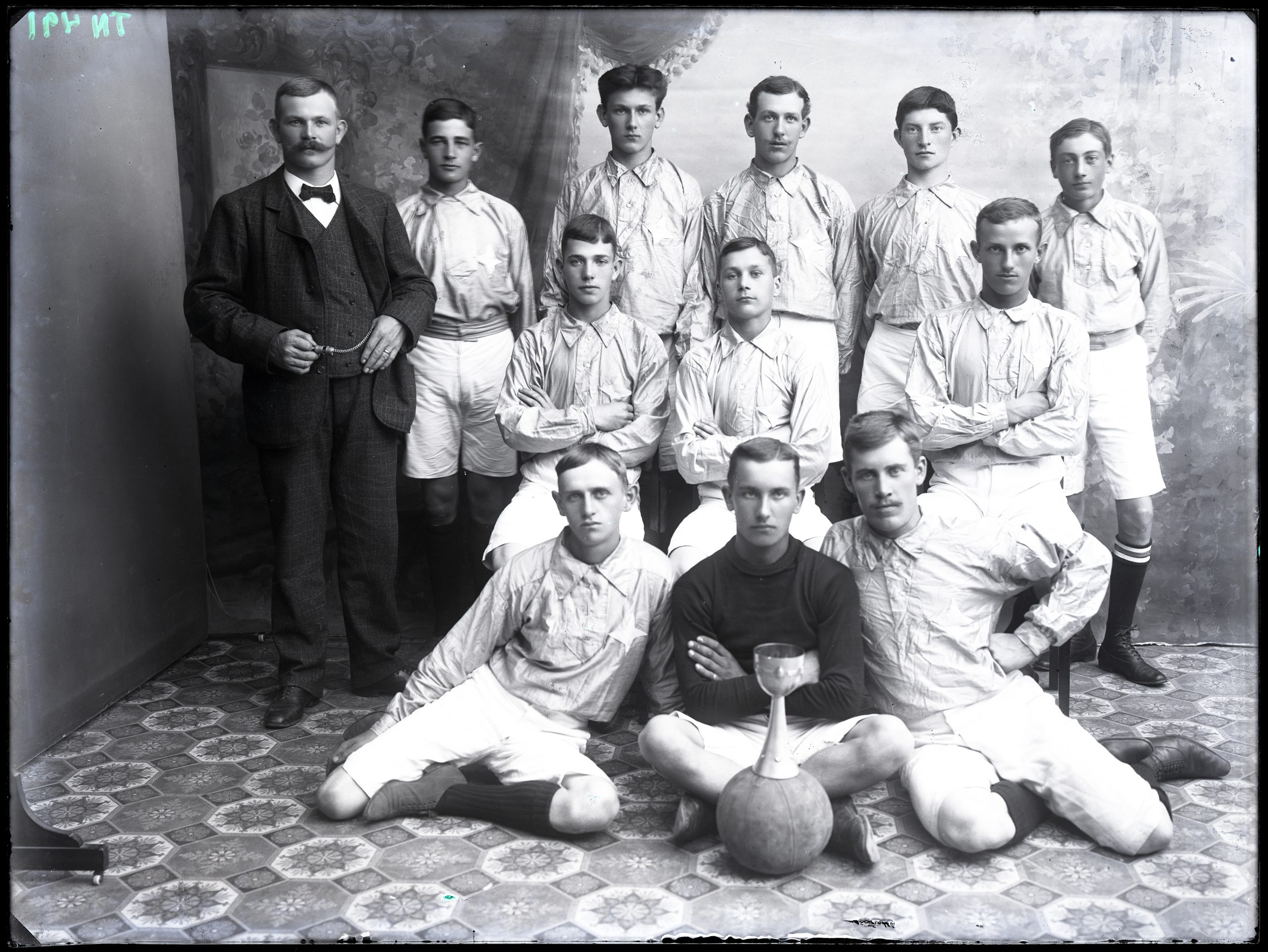 Svartvitt foto med en grupp män iförda skjortor och kortbyxor står och sitter framför en fotboll med en pokal.