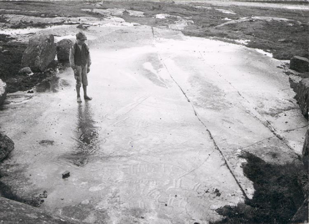  Svartvitt foto. En barfota pojke med keps och väst står på en regnvåt häll. Ristningar i hällen kan så vitt anas som skuggor. 