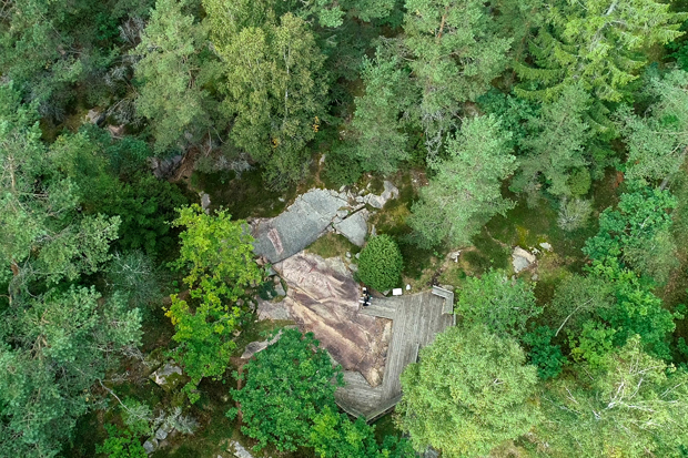 Foto. Blandskog i fågelperspektiv. I en glänta, berg i dagen mellan grön gräsmark. 