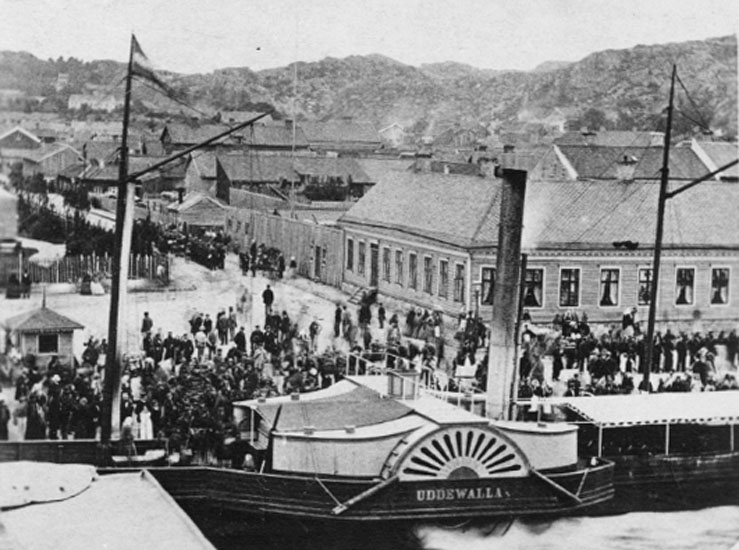 Svartvitt foto. Hjulångare med hög skorsten framför en kaj med folksamling. I bakgrunden stadsbebyggelse.