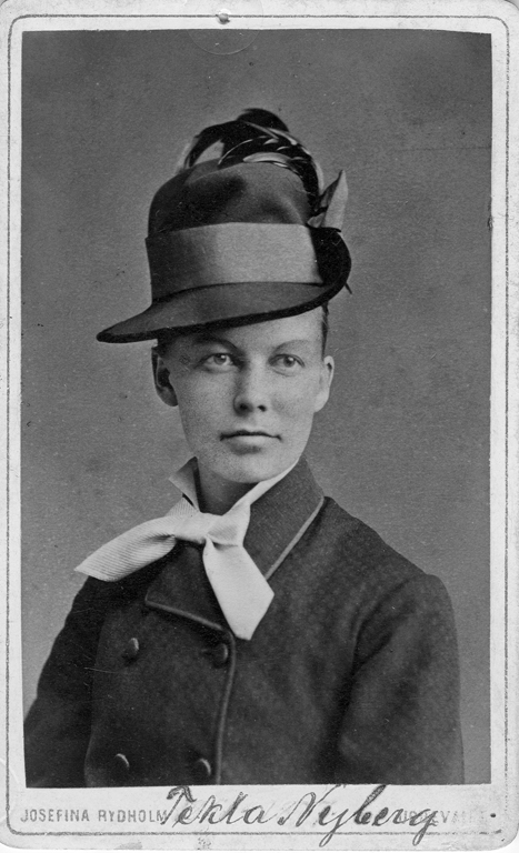 Svartvitt porträttfoto av Tekla Nyberg i 30-års-åldern. Hon bär en hög filthatt med fjäder och en dubbelknäppt mörk skepparkavaj.