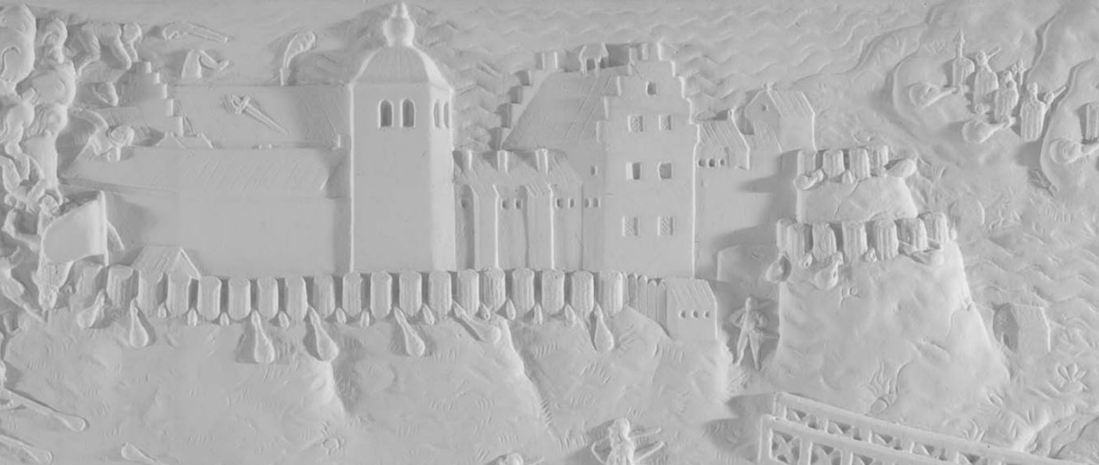 Relief i gips. På en höjd tronar en borg med torn och trappgavlar. En flagga sticker upp ur muren som omgärdar de fyra huskropparna. 