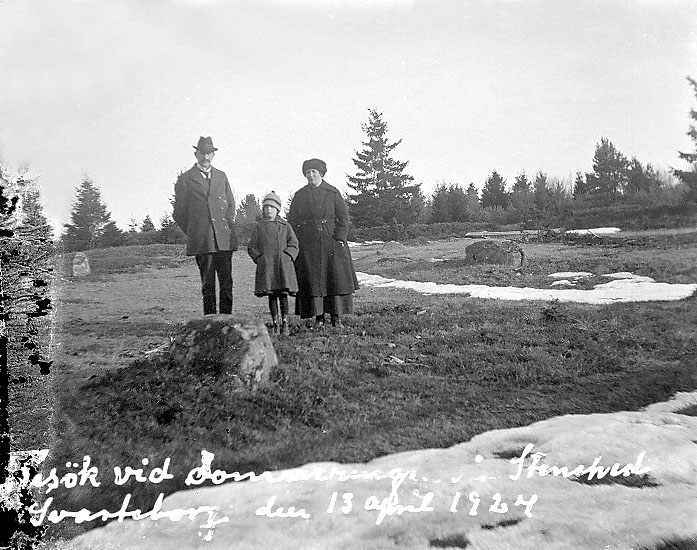 Svartvitt foto med 2 vuxna och ett barn bakom en halvmeterstor klumpsten. De står på en gräsyta med fler stenar och i bakgrunden anas skogsranden.