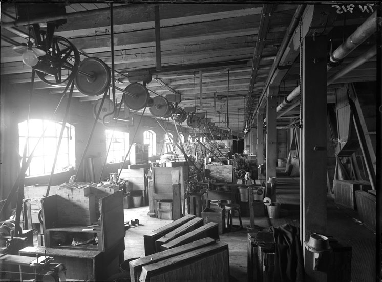 Svartvitt foto med interiör från fabrikslokal. I taket syns remdriften som driver maskinerna.