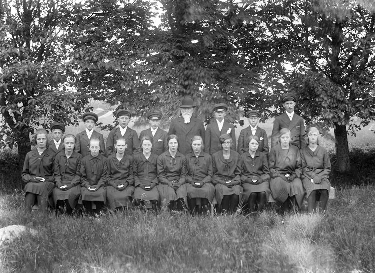 Svartvitt gruppfoto med mörklädda ungdomar som står och sitter runt en hattförsedd man med prästkrage och skägg.