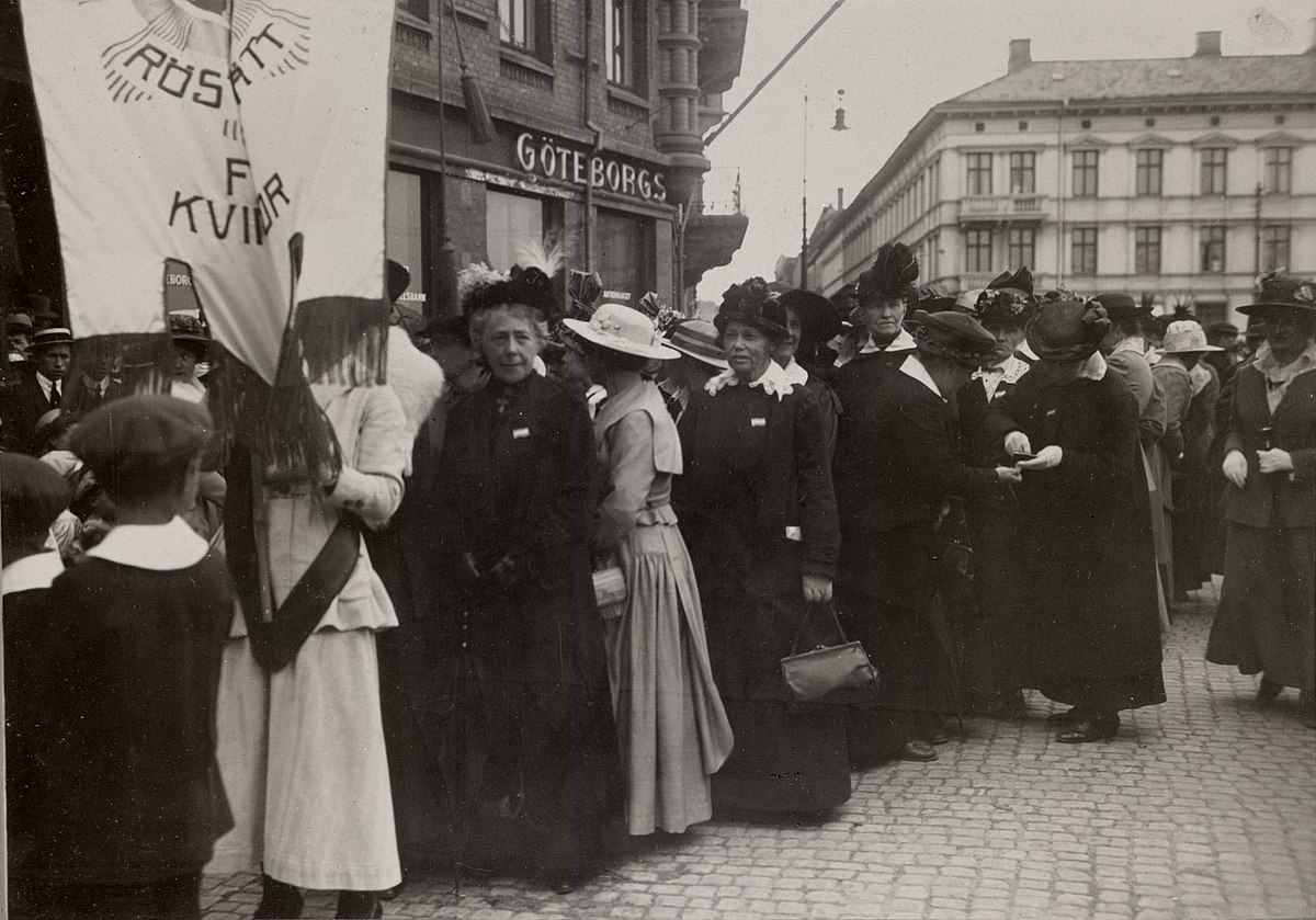 Svartvitt foto med en rad kvinnor i stor hattar och långa kjolar. Längst fram i protesttåget en kvinna med rösträttsbanderoll. 