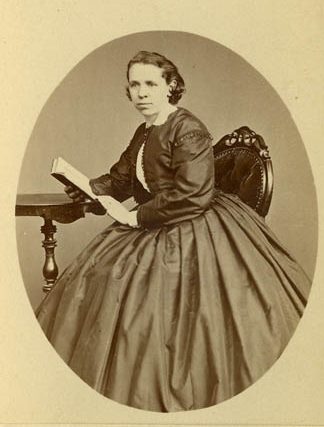 Gulnat porträttfoto av en sittande kvinna i krinolin med en bok i handen.