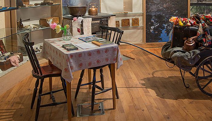 Foto. Ett köksbord för max 4 personer med två pinnstolar, duk och blommor i papper. Till höger en gammeldags trä-vagn lastad med personliga tillhörigheter.
