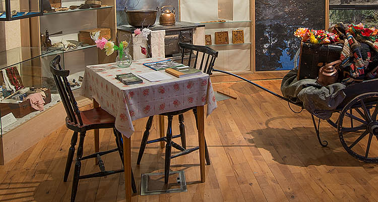Foto. Ett köksbord för max 4 personer med två pinnstolar, duk och blommor i papper. Till höger en gammeldags trä-vagn lastad med personliga tillhörigheter.
