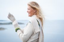 Foto. Pernille Silfverbeerg i profil vid ett stilla hav i ljus stickad tröja och halsduk. Hon har håller upp händer iklädda vantar i Bohus Stickning och ler.