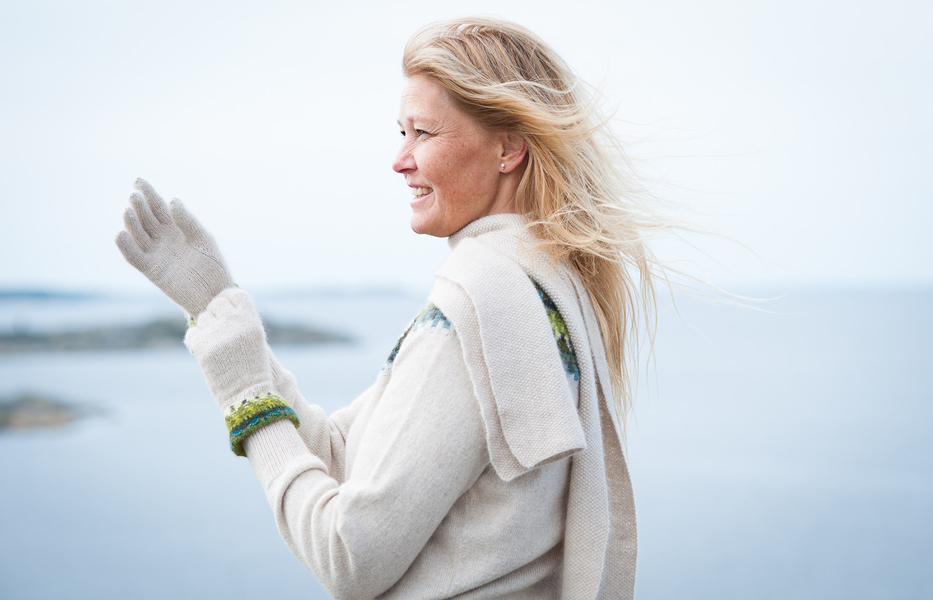 Foto. Pernille Silfverbeerg i profil vid ett stilla hav i ljus stickad tröja och halsduk. Hon har håller upp händer iklädda vantar i Bohus Stickning och ler.