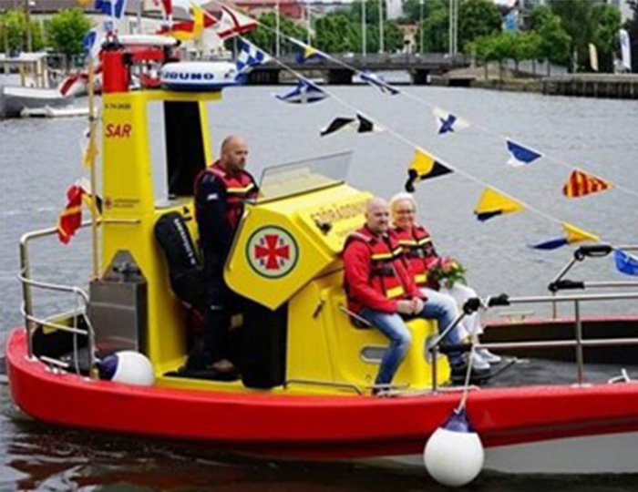En gul & röd båt i vattnet. En person kör båten och två andra sitter fram i båten. Båten pryds av ett band av flaggor.