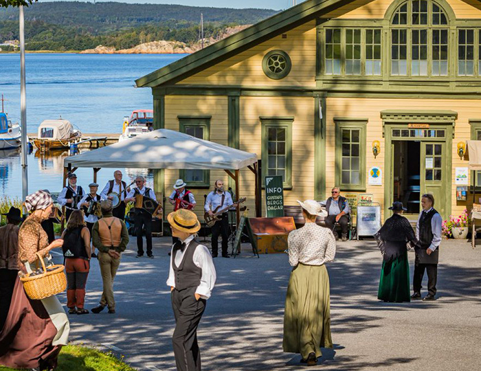 Ett myller av människor framför ett gult hus med gröna detaljer. Huset ligger vid vattnet och det står ett band med dragspel, banjo och bas och spelar.