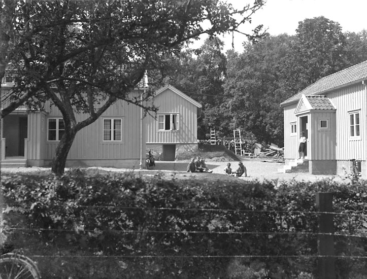 Svartvitt foto. En sommarbild. Några barn leker på gårdsplanen, omgivna av tre hus.