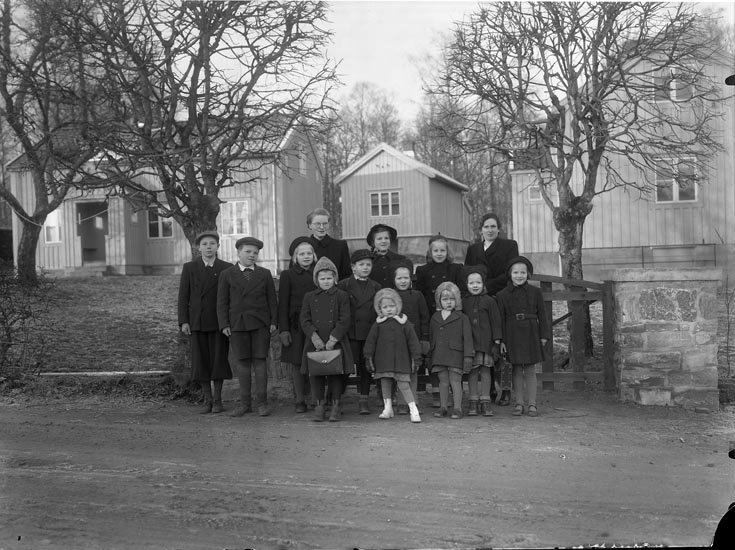 Svartvitt foto. En grupp vinterklädda barn och två vuxna står framför några byggnader.