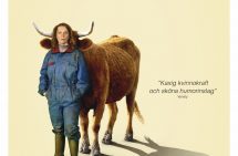En kvinna står i blåställ och stövlar, bakom henne står en brun ko coh gömmer sig bakom kvinnan.