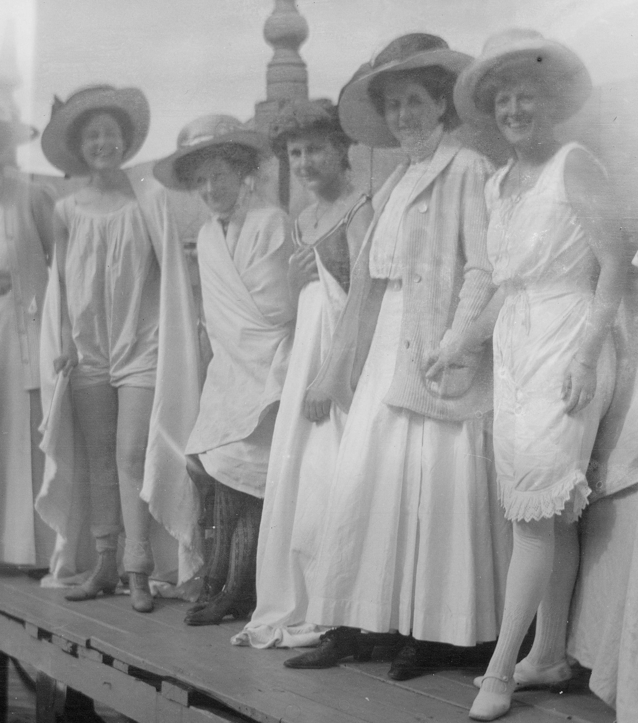 Svartvitt foto. Fem kvinnor i ljusa baddräkter, linnen och badlakan står på en brygga. Fyra har även hatt och strumpor och skor.