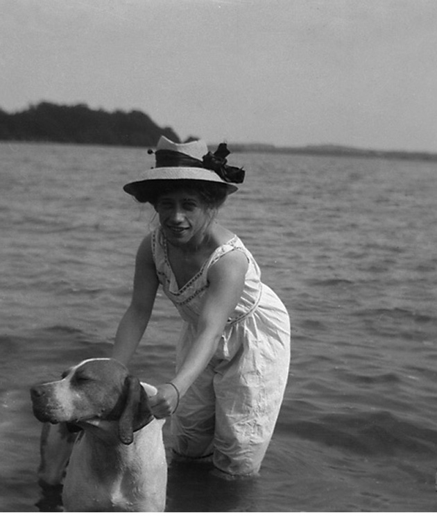 Svartvitt foto. Kvinna i ljus baddräkt och stråhatt badar tillsammans med en hund.
