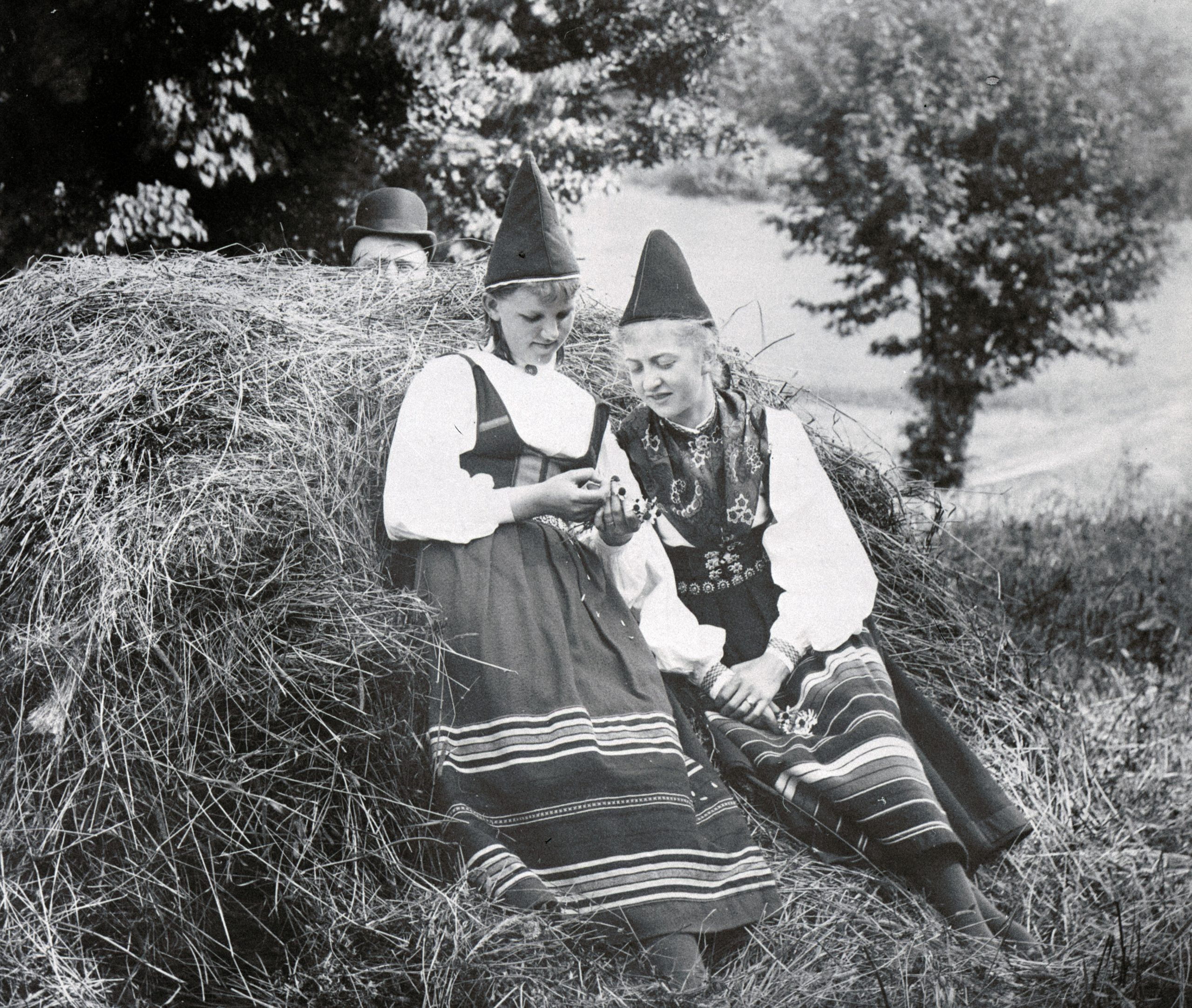 Svartvitt foto. Två unga flickor i folkdräkter med toppiga hattar vilar vid en höstack.
