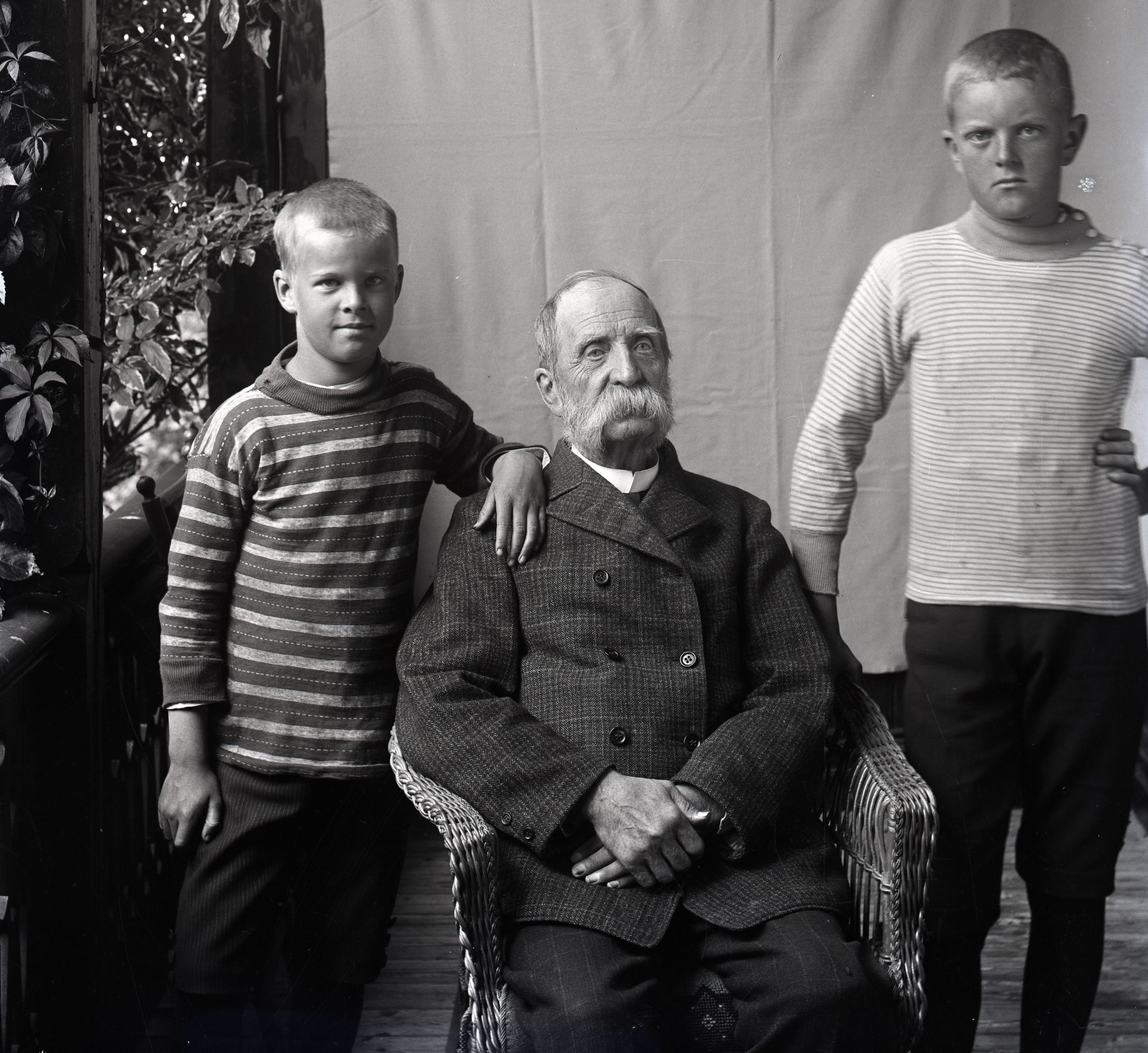 Svartvitt foto. En äldre sittande man med stor mustasch omgiven av två pojkar cirka 7 och 9 år gamla.
