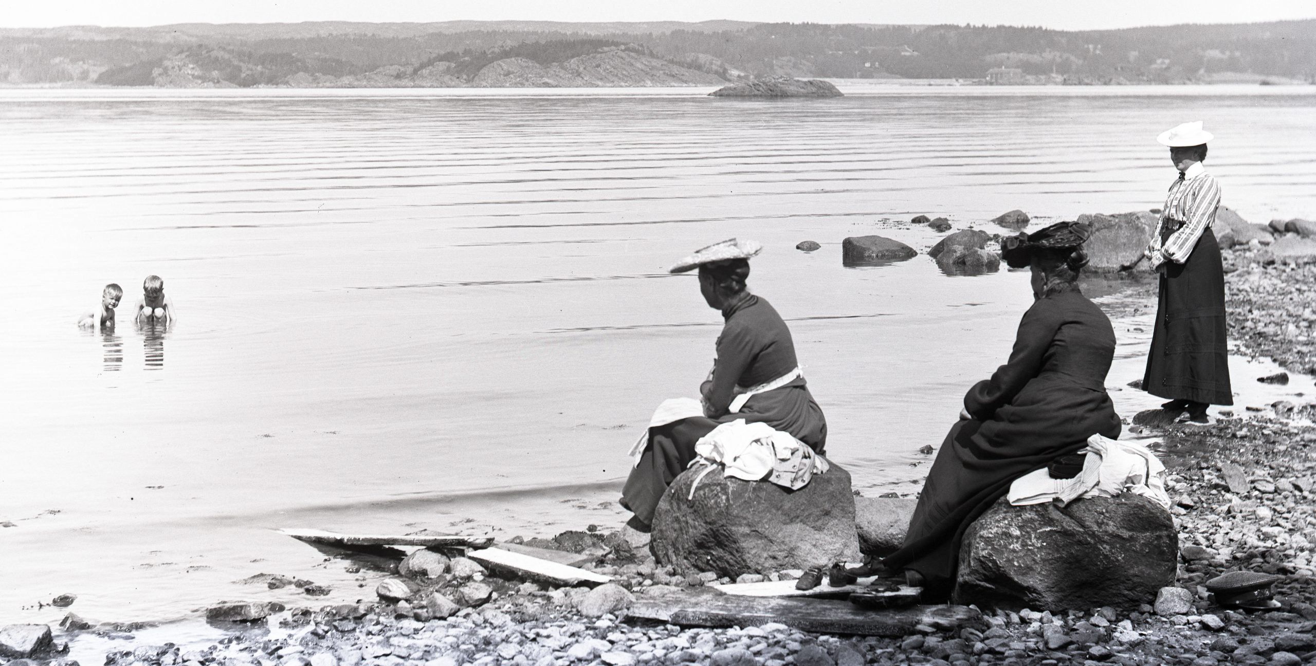 Svartvitt foto. Tre kvinnor på stranden tittar på två små pojkar som badar i havet.