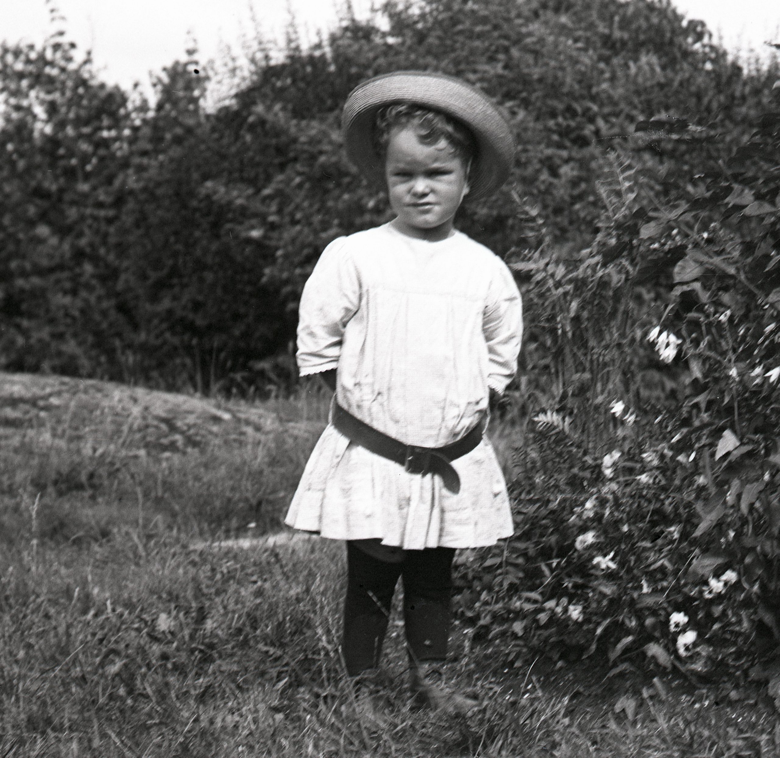 Svartvitt foto. Pojke, cirka två år, klädd i kolt och hatt står i en trädgård.
