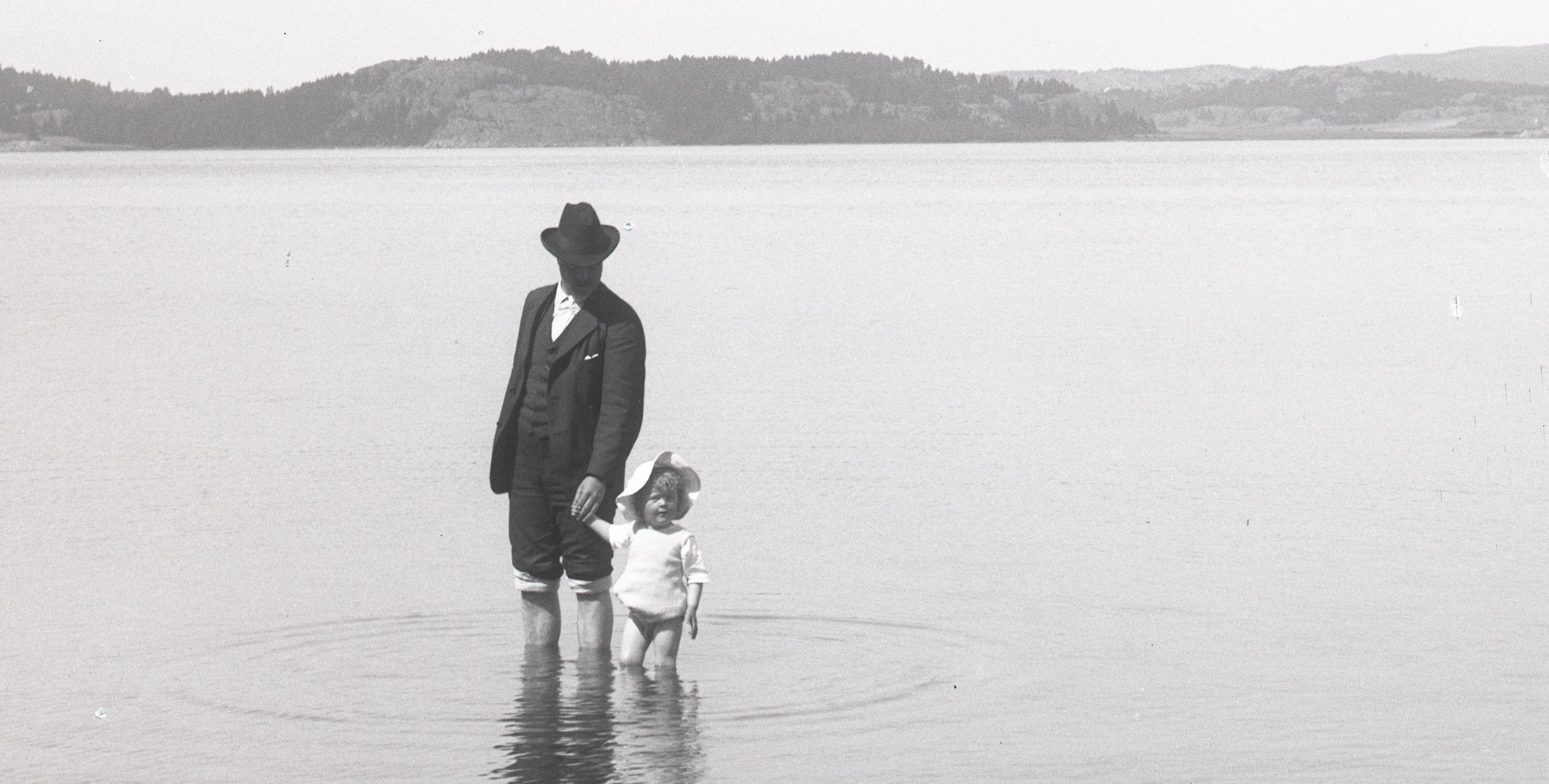 Svartvitt foto. En man i hatt, kavaj och uppkavlade byxor stå ute i vattnet med ett litet barn.
