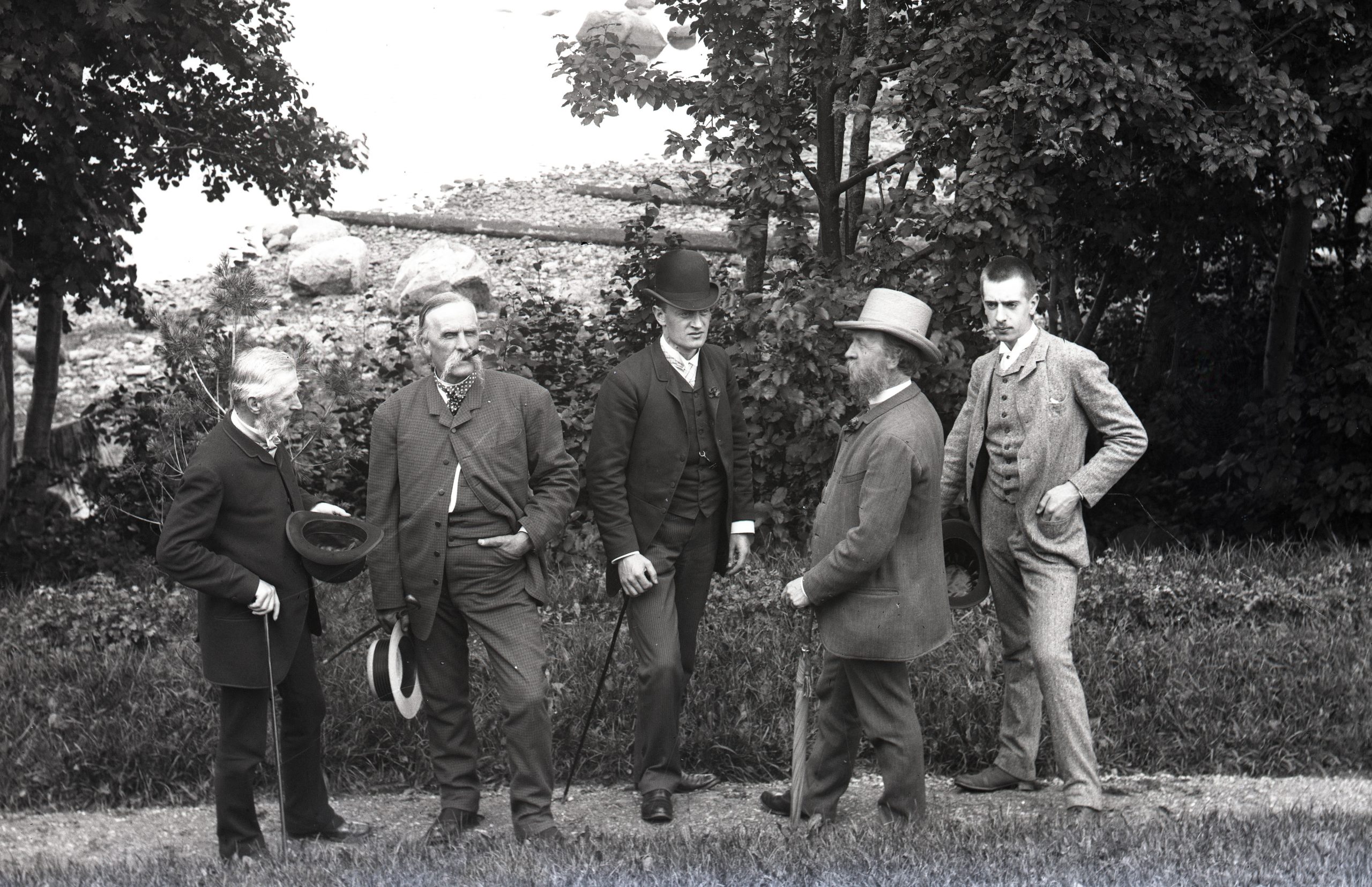 Svartvitt foto. Grupp med fem herrar i kostym på en promenadväg