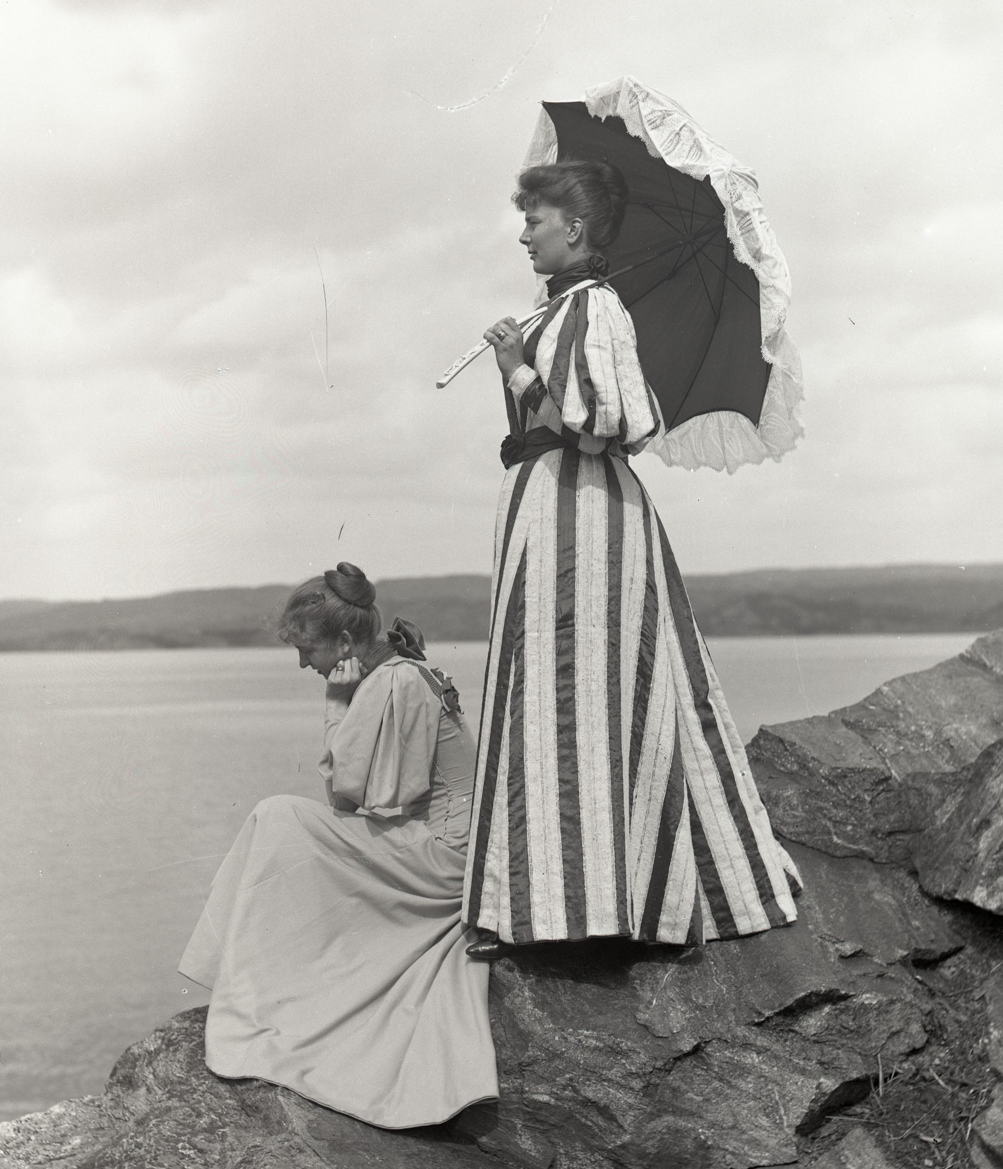 Svartvitt foto. Två unga kvinnor i profil vid havet. En av dem sitter den andra står upp med ett parasoll.