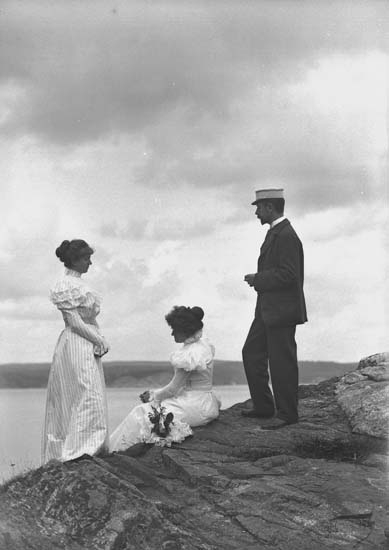 Svartvitt foto. Två unga kvinnor i ljusa dräkter och en man i mörk kostym blickar ut över havet.