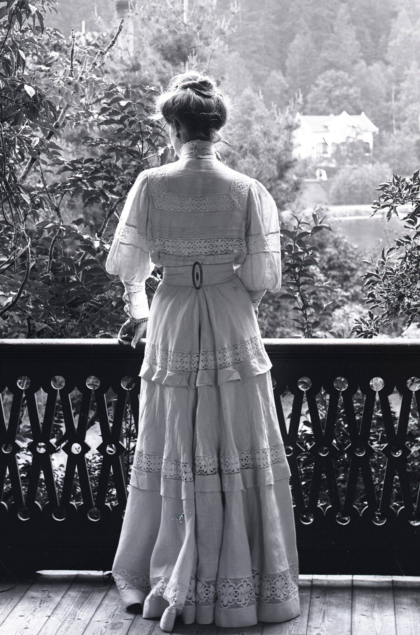 Svarvitt foto. Kvinna sedd bakifrån, står i en luftig lång dräkt på en veranda.